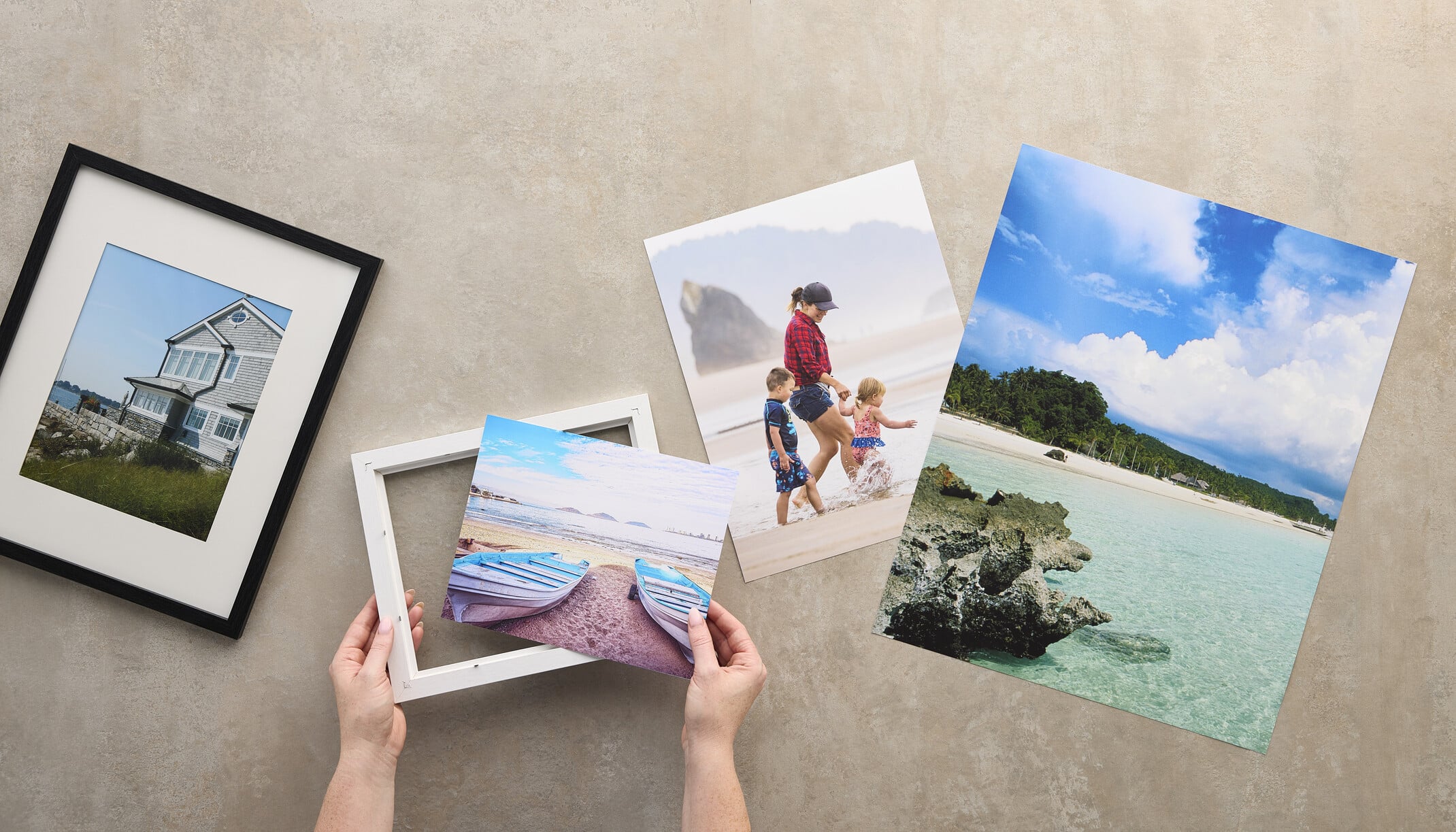 A person's hands hold a white picture frame over a print of two boats on a beach, surrounded by other framed and unframed landscape and family photos on a gray surface.