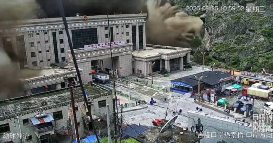 A large cloud of dust and debris billows from a mountain slope behind a multi-story government building at a border crossing.