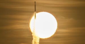A rocket ascends vertically, silhouetted against a large, bright sun in a hazy sky.