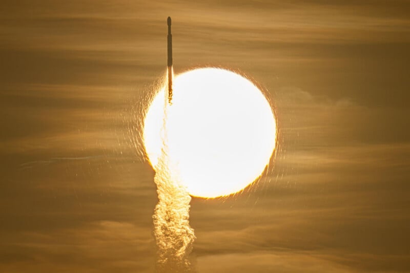 A rocket ascends vertically, silhouetted against the bright, golden disk of the sun in a hazy sky.