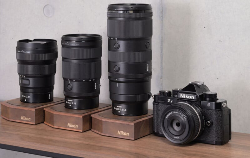 Three Nikon lenses on wooden stands sit next to a Nikon Zf camera with a 40mm lens on a wooden surface.