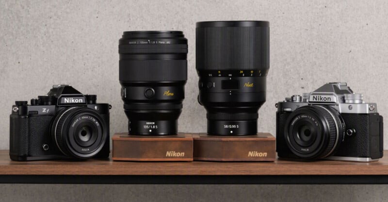 Two Nikon Zf cameras, one black and one silver, are positioned on a wooden shelf alongside two large Nikon lenses, the 135mm Plena and 58mm Noct, each resting on a branded wooden stand.