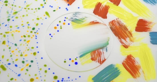 A circular glass pane sits on a white surface, featuring colorful paint splatters on the left and bold, textured brushstrokes in yellow, orange, and blue on the right.