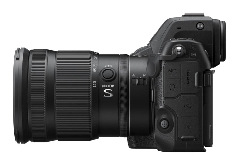 A side view of a black mirrorless camera with a zoom lens attached, showing the lens focus ring, function button, and side ports for microphone, headphones, and USB-C.