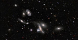 A deep-space view showing a dense cluster of various spiral and elliptical galaxies scattered across a dark, star-filled background.