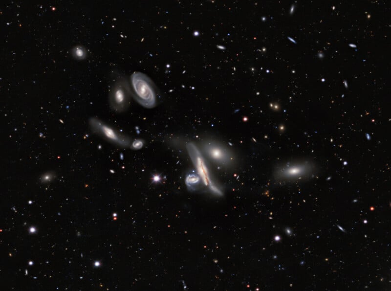 A deep-space view showing a dense cluster of various spiral and elliptical galaxies scattered across a dark, star-filled background.