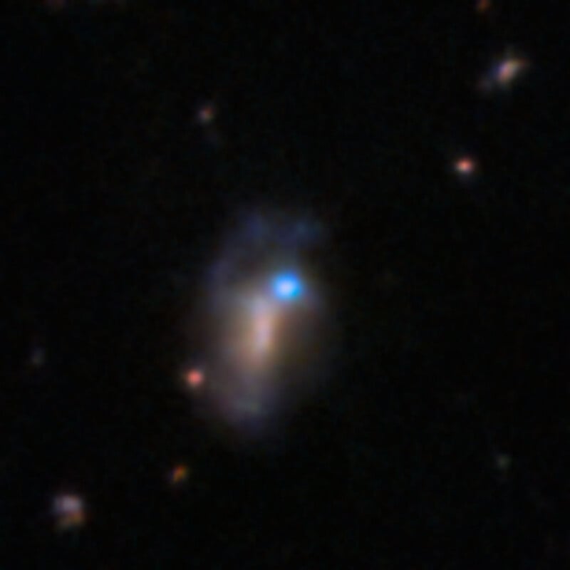 A distant, irregular galaxy with a bright blue core and faint, diffuse reddish-brown structures against a black background.