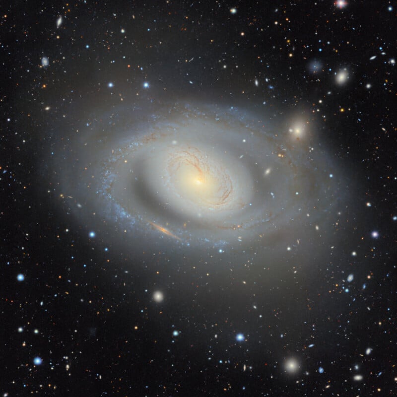 A large, luminous spiral galaxy with a bright central core and faint, swirling blue arms sits against a backdrop of numerous distant stars and smaller galaxies.