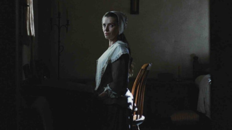 A woman in a historical white cap and shawl stands in a dimly lit room, looking back over her shoulder toward the camera.