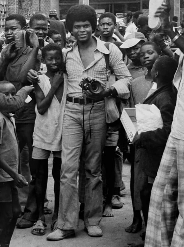 A young man with an afro stands in a crowd of children, holding a camera and smiling while wearing a plaid shirt and light-colored pants.