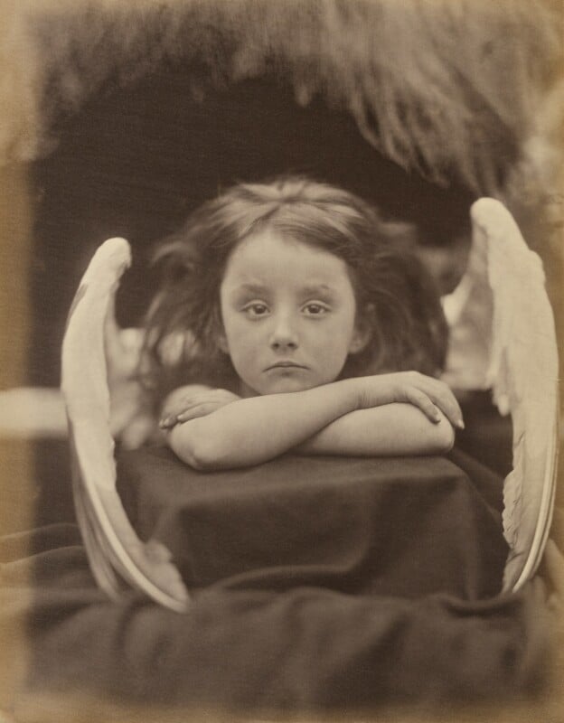 A sepia-toned portrait of a young child with long, dark hair and feathered wings, resting their chin on crossed arms.