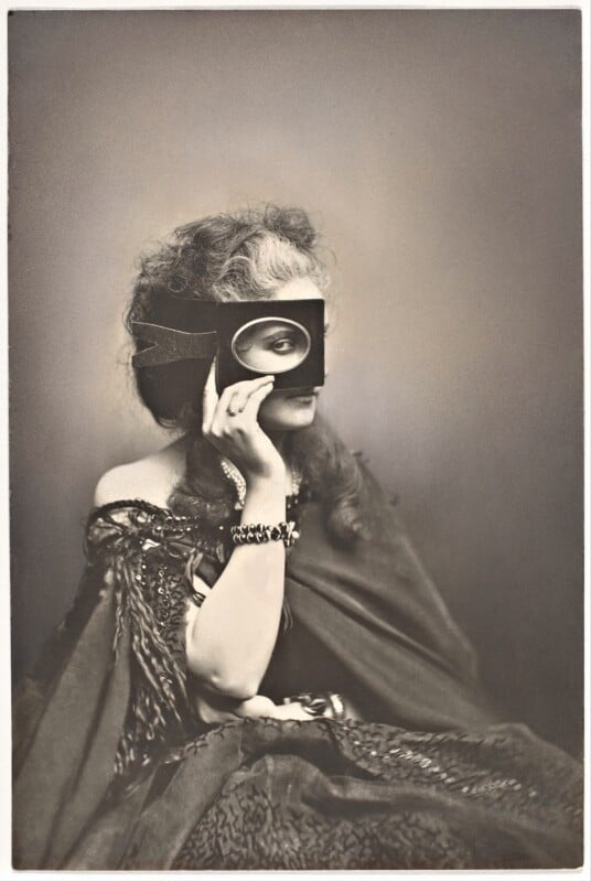 A woman with wavy hair wears an ornate dark dress and holds a rectangular frame with an oval cutout over one eye, revealing her gaze.