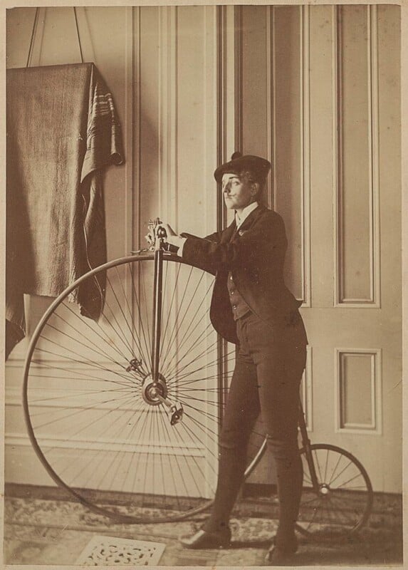A person in a suit and cap stands beside a large-wheeled penny-farthing bicycle indoors against a paneled wall.