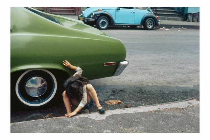 A young girl crouches in the street next to a green car, reaching toward the ground near the rear tire, with a blue Volkswagen Beetle parked in the background.