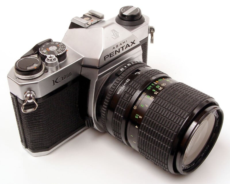 A silver Asahi Pentax K1000 35mm film SLR camera with a black 35-70mm zoom lens attached, viewed from a high angle against a white background.