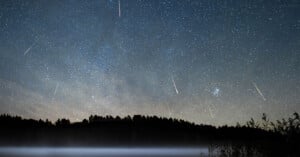 A night sky filled with stars and several bright meteor streaks above a dark, silhouetted forest and a misty lake.