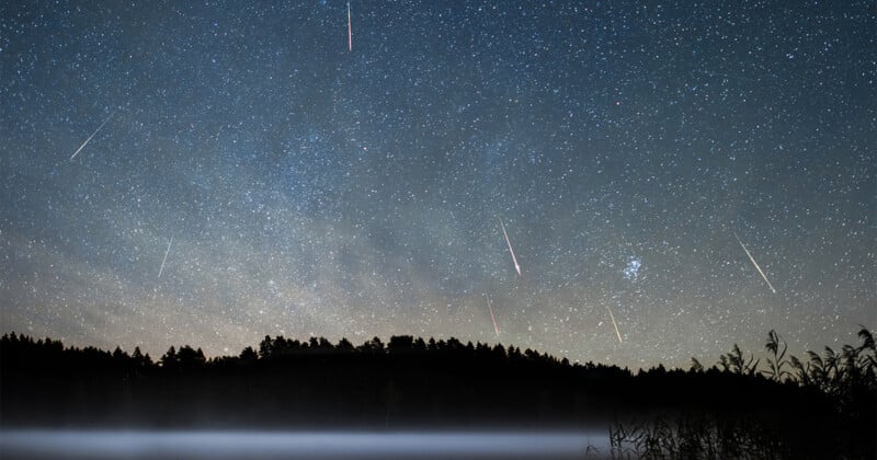 A night sky filled with stars and several bright meteor streaks above a dark, silhouetted forest and a misty lake.