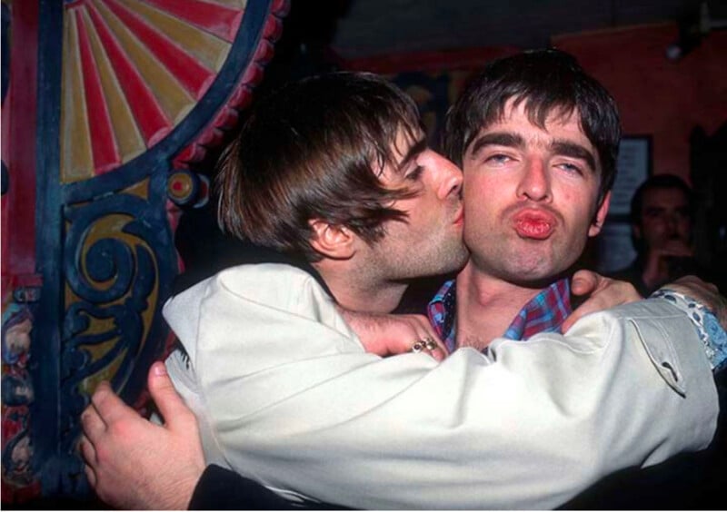 Liam Gallagher kisses his brother Noel Gallagher on the cheek while they embrace in a dimly lit room.