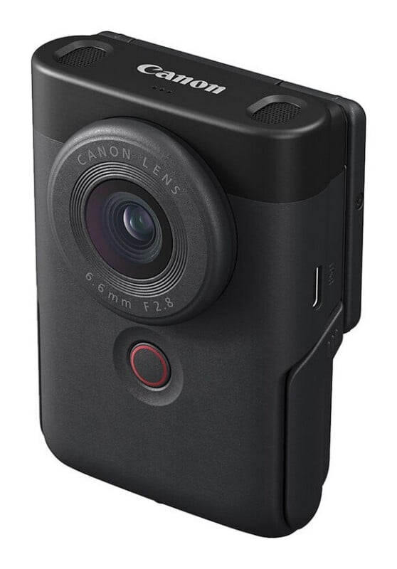 A black Canon PowerShot V10 vlogging camera shown from a front-facing angle, featuring a prominent lens, a circular record button, and two top-mounted microphones.