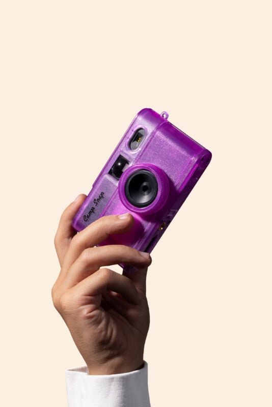 A hand holds a translucent, glittery purple Camp Snap digital camera against a plain, light-colored background.