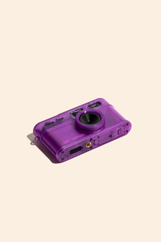 A purple, translucent digital camera with a lens and "Camp Snap" branding sits at an angle on a light beige background.
