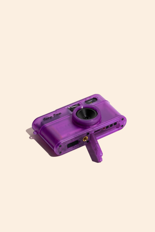 A translucent purple digital camera with a lens, viewfinder, and open battery compartment sits on a light beige background.