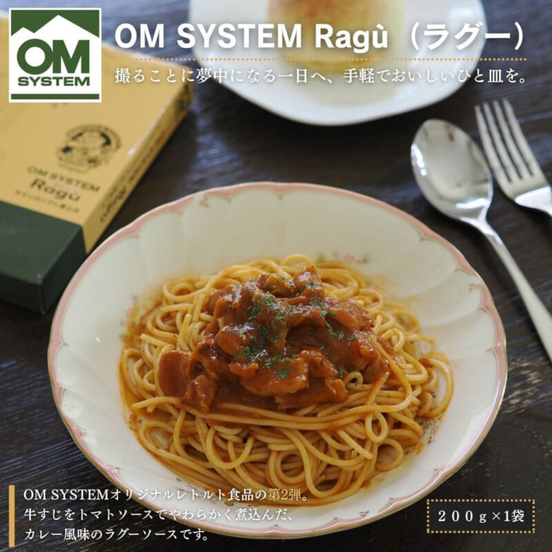 A plate of spaghetti topped with a beef ragu sauce, accompanied by a fork, a spoon, and a box of OM SYSTEM Ragù on a dark wooden table.