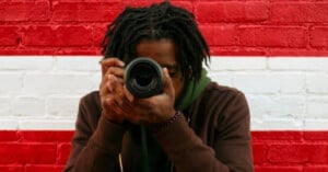 A person with dreadlocks holds a camera up to their face, obscuring their eyes, against a background of red and white bricks.