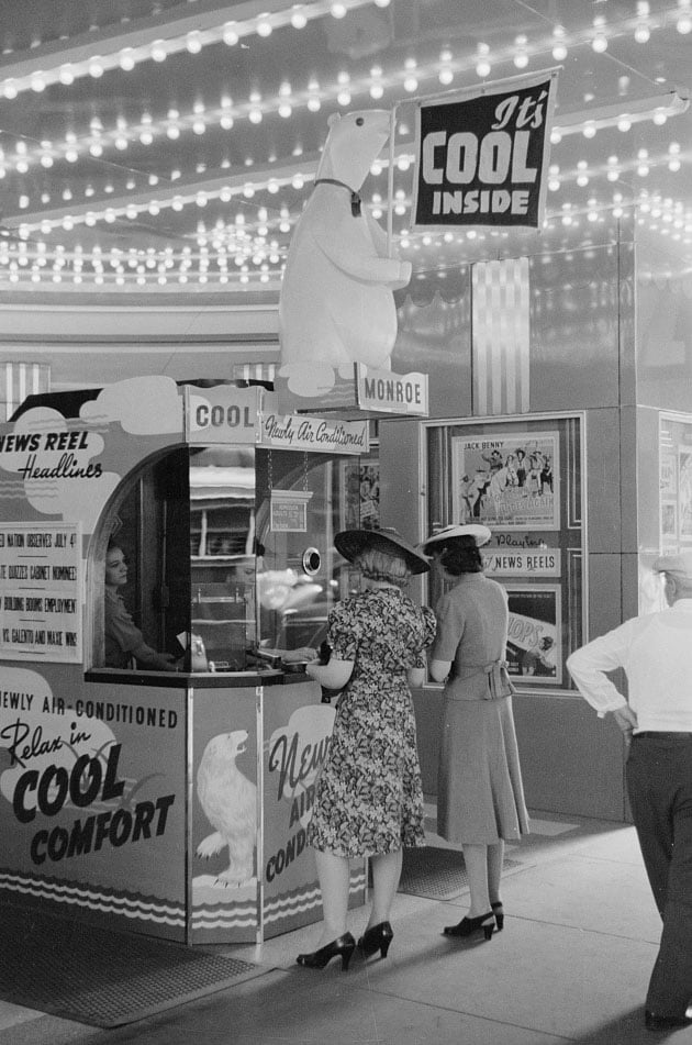 Two women stand at a movie theater ticket booth featuring a polar bear statue and signs advertising air conditioning.