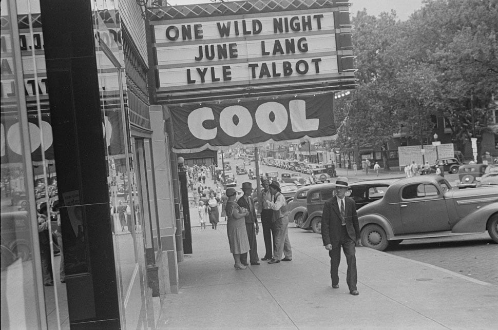 A street scene in front of a movie theater with a marquee advertising the film "One Wild Night" starring June Lang and Lyle Talbot, with a "COOL" banner hanging below and pedestrians walking on the sidewalk.