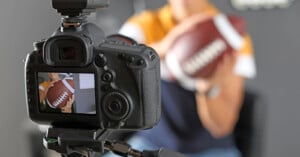A camera on a tripod faces a person holding an American football, with the camera's screen displaying a live view of the person and the ball.