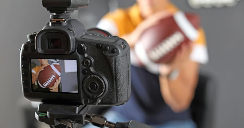 A camera on a tripod faces a person holding an American football, with the camera's screen displaying a live view of the person and the ball.