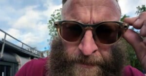 A close-up shot of a man with a thick beard wearing translucent-framed sunglasses, holding the frame with his hand.