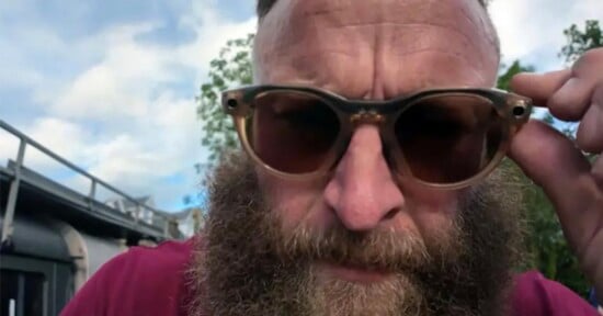 A close-up shot of a man with a thick beard wearing translucent-framed sunglasses, holding the frame with his hand.