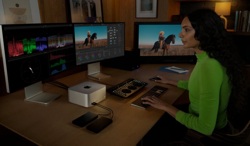 A person in a bright green long-sleeved shirt sits at a desk working on a computer with three monitors, a keyboard, and a color grading control panel.