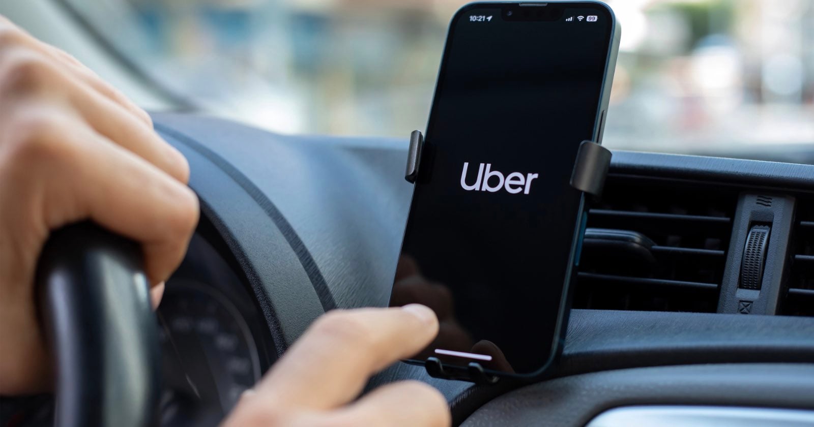 A driver's hands are visible on a steering wheel and touching a smartphone mounted on a car dashboard displaying the Uber logo.