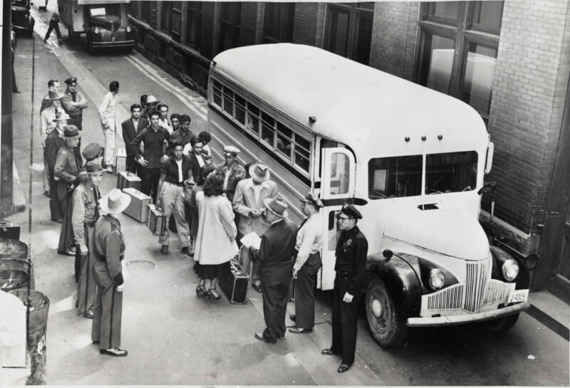 A group of men and one woman stand in a line next to a bus in an alleyway, supervised by several uniformed officers.