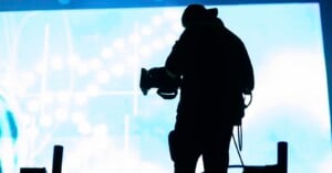 A silhouetted videographer stands in front of a bright, glowing blue screen while operating a professional video camera.