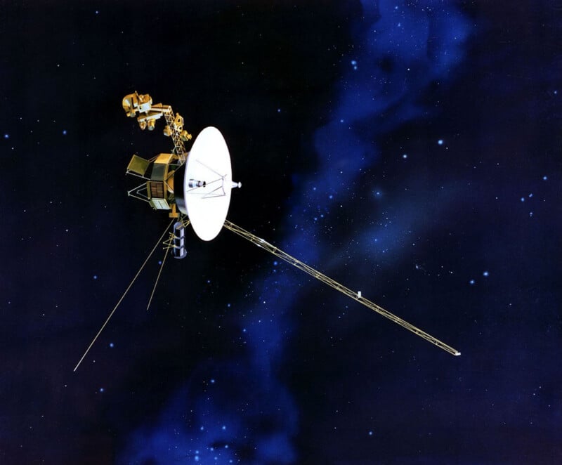 A Voyager space probe floats in deep space against a backdrop of stars and a blue nebula.