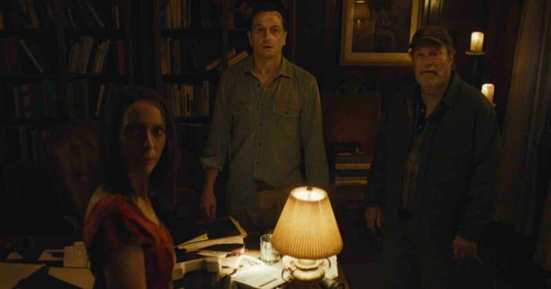 Three people stand in a dimly lit, wood-paneled room with bookshelves, illuminated by a table lamp in the foreground.