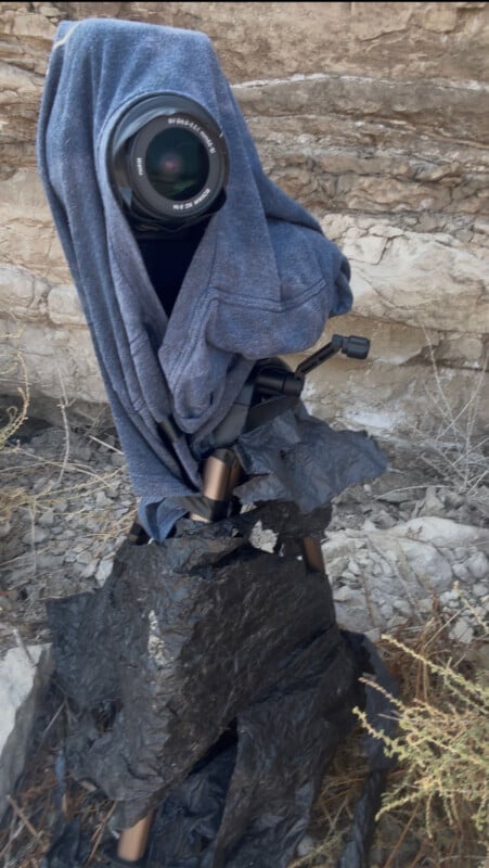 A camera on a tripod is draped with a blue shirt and partially wrapped in black plastic, positioned against a rocky cliff face.