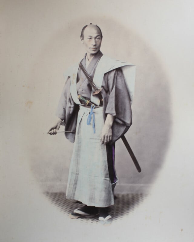 A full-length portrait of a Japanese man in traditional samurai attire, including a kimono, hakama, and two swords tucked into his sash.