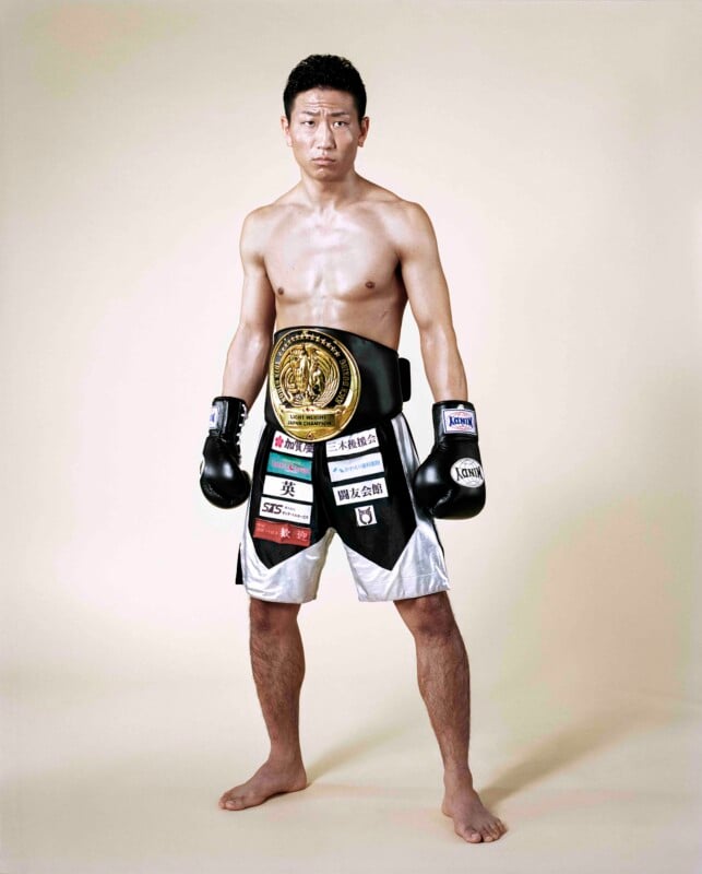 A shirtless male kickboxer stands barefoot against a plain background, wearing black and silver trunks with sponsor patches and a championship belt.