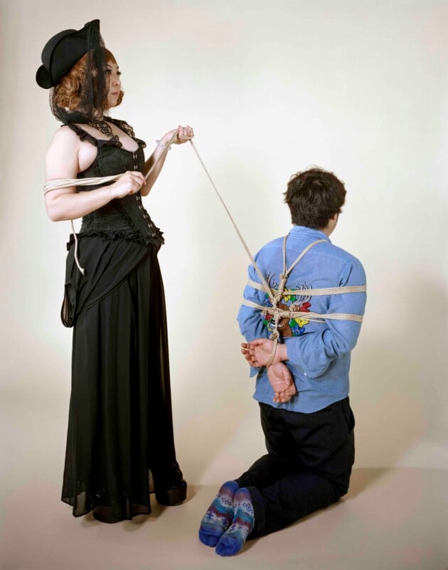 A woman in a black corset and hat stands holding a rope that is tied around a kneeling man wearing a blue shirt and black pants.