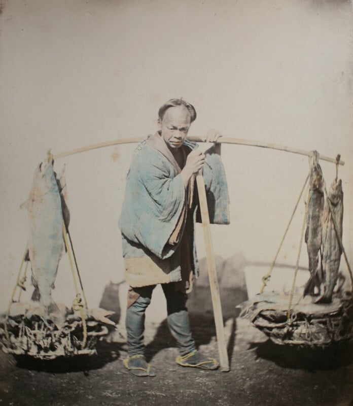 A man in a blue kimono carries a bamboo pole across his shoulders with baskets of fish hanging from each end.