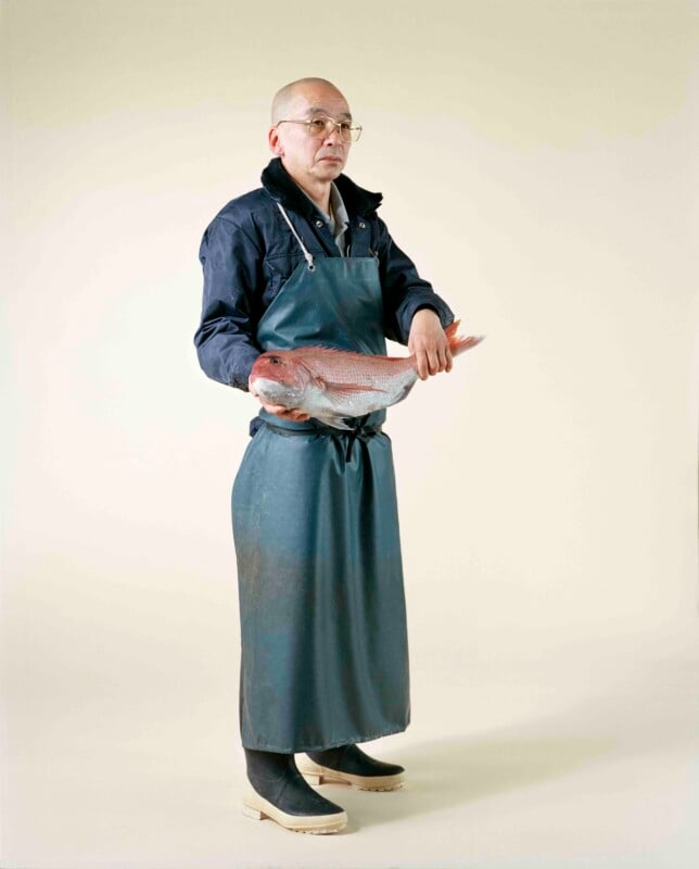 A bald man wearing glasses, a dark jacket, and a long green apron stands against a plain background, holding a large red fish.