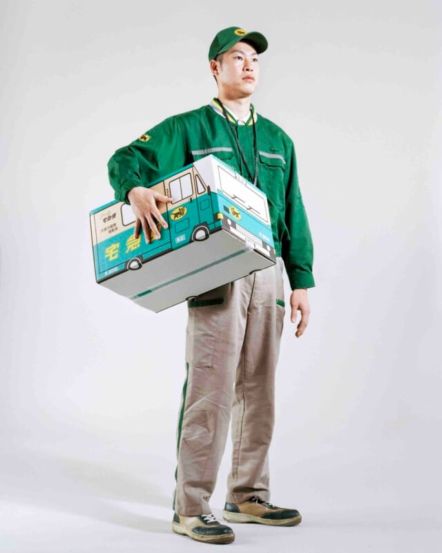 A delivery worker in a green uniform and cap stands against a white background, holding a large cardboard box printed with the design of a delivery truck.