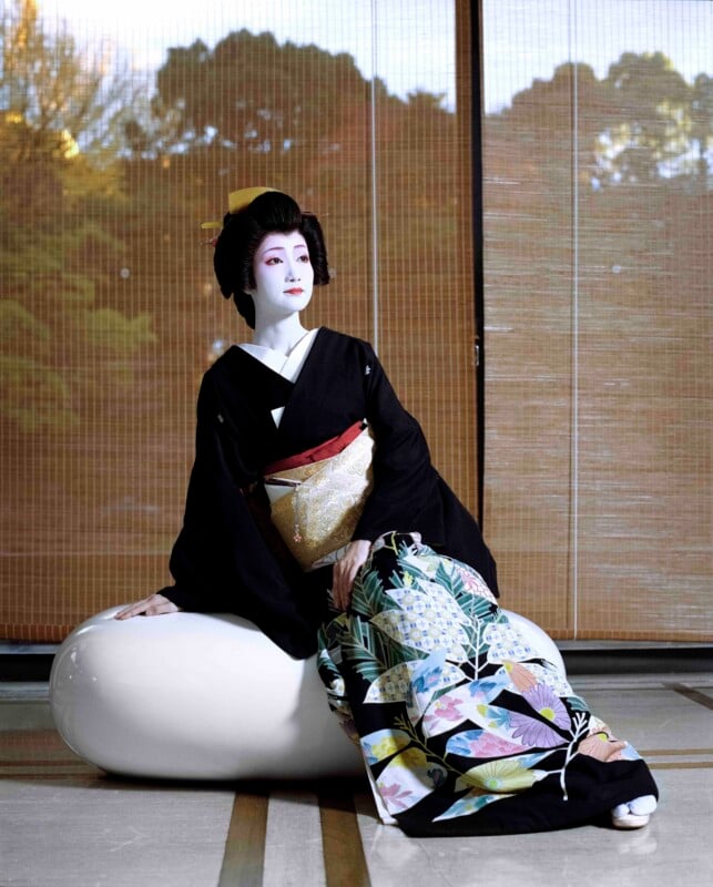 A woman in traditional geisha makeup and a black kimono with a floral skirt sits on a large, smooth white oval object in front of bamboo blinds.