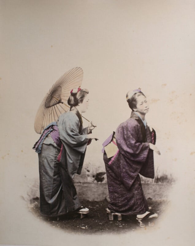Two women in traditional Japanese kimono and wooden geta sandals stand on a dirt path, one holding a large paper umbrella.