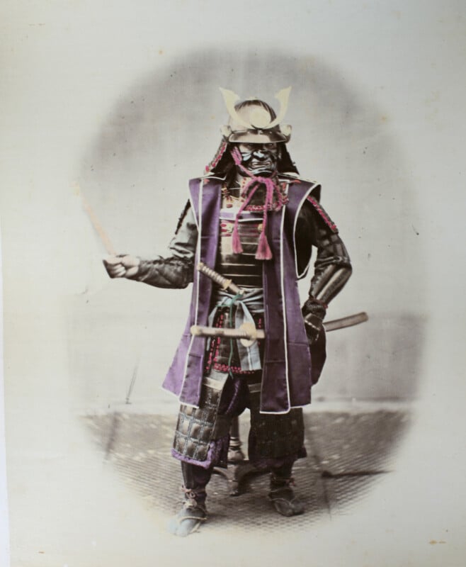 A hand-colored studio portrait of a samurai in full armor, wearing a horned helmet and a fierce mask, holding a baton.